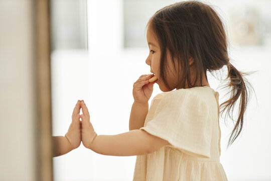 Side view of Asian little girl eating cookies and looking at her reflection in the mirror at home