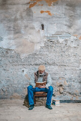 Fototapeta na wymiar Old man with gray beard reads a book on a chair against an old concrete wall
