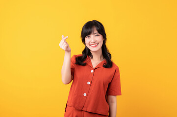 Portrait young beautiful asian woman happy smile dressed in orange clothes showing mini heart hand gesture isolated on yellow studio background.