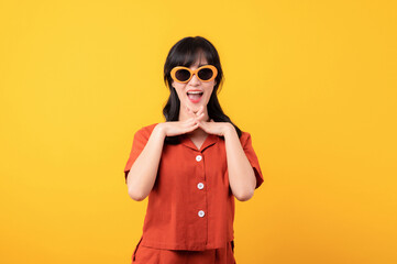 Portrait young beautiful asian woman happy smile dressed in orange clothes and sunglasses looking celebrate with hand on her face isolated on yellow studio background.