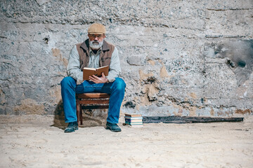 Fototapeta premium An old man sits on a chair near an old concrete wall. Reading book