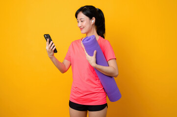Naklejka premium Portrait young asian sports fitness woman happy smile wearing pink sportswear and smartphone doing exercise training workout against yellow studio background. technology wellness lifestyle concept.