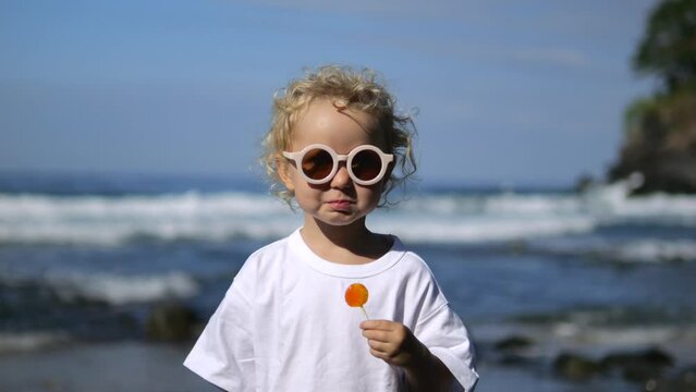 Slow motion, portrait of little girl in cool hipster round glasses. Little Caucasian stylish fashionable girl eats lollipop while on sea or ocean. Baby on sea basks in warm sun and eats lollipop.