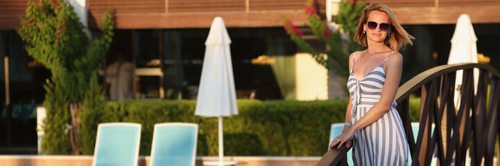 Beautiful woman in summer sundress on territory of hotel by pool at sunset. Summer vacation and travel concept