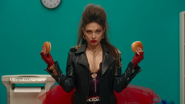 A freak woman stands in the office holding a hamburger in her crossed arms. The woman takes turns greedily taking bites from both burgers, stuffing her mouth full. Advertising. Slow motion.