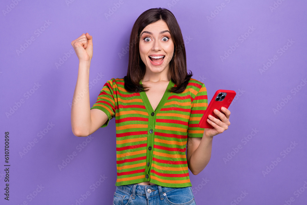 Sticker Portrait of optimistic impressed girl dressed striped cardigan hold smartphone raising fist win lottery isolated on violet background