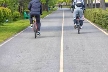 Two cyclists ride along an asphalt path through a green summer park.