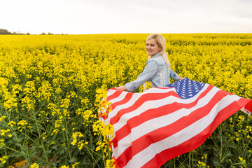 Fototapeta attractive woman holding an American flag in the wind in a field of rapeseed. Summer landscape against the blue sky. Horizontal orientation. obraz