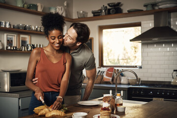 Interracial couple, kiss and hug in kitchen for cooking, morning breakfast, love or caring relationship at home. Man kissing woman preparing food, meal or cutting ingredients on table at the house