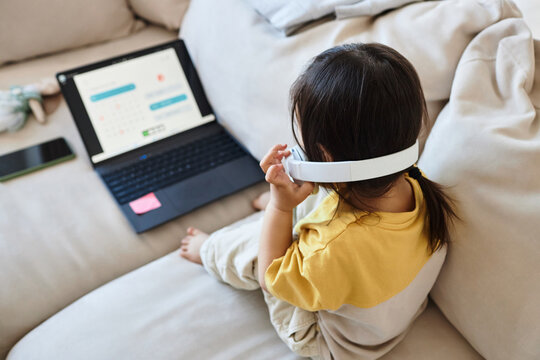 Rear view of little girl in wireless headphones sitting on sofa in front of laptop and watching cartoons