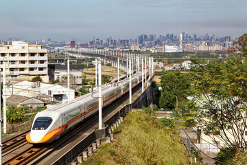 
A train of Taiwan High Speed Rail (THSR) traveling through a railway curve in the countryside with modern skyscrapers of Taichung City in the distant background under blue sunny sky