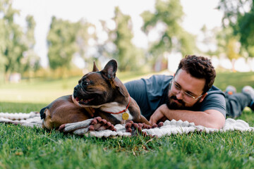 bearded man resting in the park with his pet dog french bulldog outdoors lying on a sheet summer...