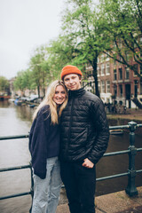 Smiling young couple on bridge over rainy canal in Amsterdam boats, trees, water, sweaters, hats, jeans, 20's