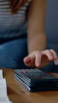 Vertical video. Close up view of young homeowner woman sitting on sofa using calculator to calculate monthly budget, household expenses, mortgage, rental fees, paying bills online.