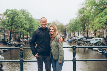Smiling middle age couple on bridge over rainy canal in Amsterdam boats, trees, water, sweaters, jeans, 40's