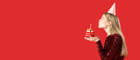 Beautiful young woman blowing out candle on tasty birthday cake against red background with space...