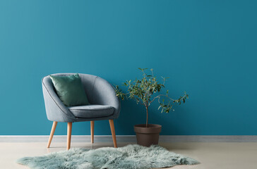 Cozy armchair with cushion, fur rug and houseplant on stool near blue wall