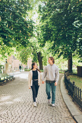 young couple walking down a tree lined street in a park holding hands and looking and smiling at each other
