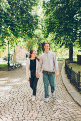 young couple walking down a tree lined street in a park holding hands and looking and smiling at each other