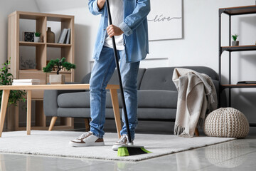 Young man sweeping carpet with broom at home