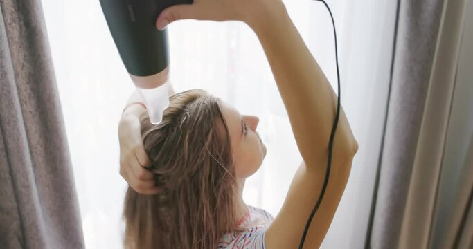 A young woman dries her long hair with an electric hair dryer.