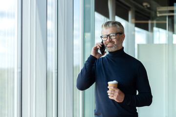 Portrait of a senior handsome gray-haired man designer, architect, engineer standing in the office by the window talking on the phone and holding a cup of coffee. Smiling looking at the camera.