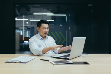 Fototapeta na wymiar Online business meeting with the team, partners. A young Asian businessman is sitting in the office at a laptop and talking on a video call, holding a remote conference.