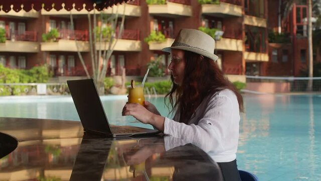 Freelancer woman working on laptop sitting in pool bar counter drinking cocktail while vacation recreation. Resting relaxing outdoors. Travel tourism, summer holidays. Freelance, distant job work.