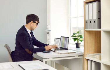 Professional financial accountant working in the office. Man in a suit and glasses sitting at his desk in front of laptop computers and looking at figures in digital business spreadsheets on screens