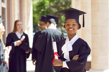 Young smiling african american girl student in a university graduate gown and diploma in her hands. Woman with crossed arms outdoor on the background of classmates. Graduation and education concept.
