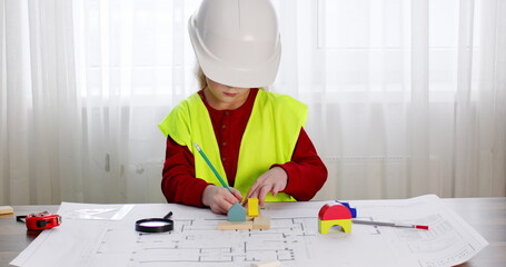 Junior schoolgirl imitates role of engineer and draws layout of building in light premise. Girl in white helmet designs on drawing with ruler and pencil