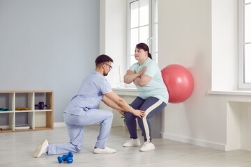 Male nurse, caregiver or health care worker helping fat overweight woman doing sport exercises with fit ball. Man physiotherapist helping plump girl in rehab. Physiotherapy and nursing help concept.