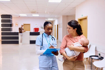 Black nurse writes data in medical record while talking to mother with baby at doctor's office.