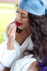 Fototapeta premium Beautiful young woman in rustic outfit with blue headscarf sitting outdoors with a large wicker basket full of fresh wildflowers