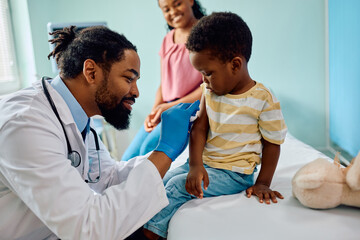 Caring African American pediatrician preparing arm of small boy for vaccination at doctor's office.