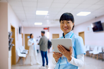 Happy Japanese nurse with digital tablet in hallway at medical clinic looking at camera.