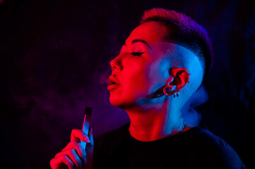 Fototapeta premium Portrait asian woman with shaved temples smokes vape in neon light.