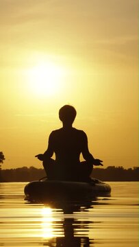 Vertical Screen: Silhouette of woman sitting in lotus pose on SUP board and meditating on water, sun setting on background. Relaxing and enjoying harmony of nature. Concept of recreation