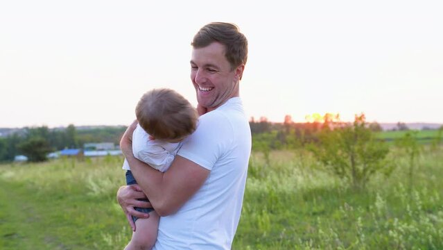 Happy man holding son in arms and playing with him outdoor in summer at sunset