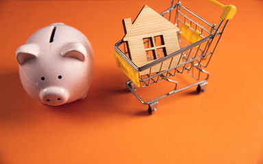 piggy bank with shopping cart and wooden house in orange background