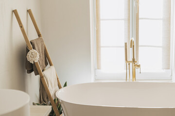 Stylish bathroom interior. Modern bathtub with golden faucet from floor, wooden ladder with towels...