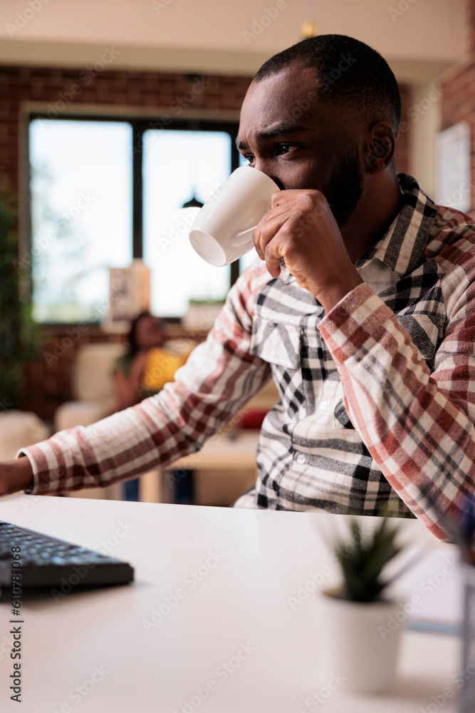 Poster african american programmer drinking coffee or tea while reading emails on computer screen in the mo