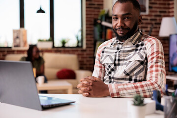 Portrait of african american male entrepreneur working remote from home smiling at camera while girlfriend is relaxing on couch. Casual man posing confident in front of laptop computer at home.