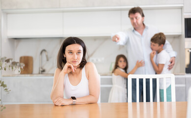 Offended mother sitting at the kitchen table, her husband, son and little daughter standing behind...