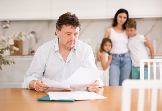 Wistful Father Looking Through Papers Sitting In The Kitchen, His Wife And Children Looking At Him With Compassion Standing Behind