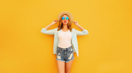 Stylish caucasian blonde young woman posing wearing straw hat, shorts and shirt on yellow background