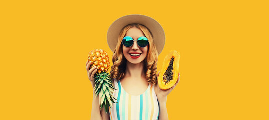 Summer portrait of happy cheerful smiling young woman with pineapple and papaya wearing straw hat, sunglasses on yellow background