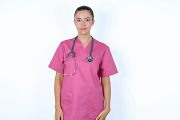 Joyful young caucasian doctor woman wearing pink uniform over white background looking to the camera, thinking about something. Both arms down, neutral facial expression.