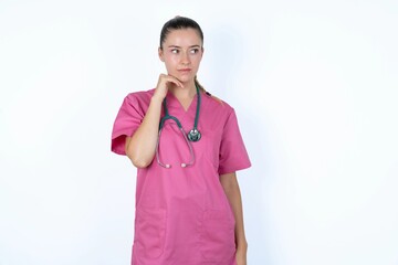 Dreamy young caucasian doctor woman wearing pink uniform over white background with pleasant expression, looks sideways, keeps hand under chin, thinks about something pleasant.