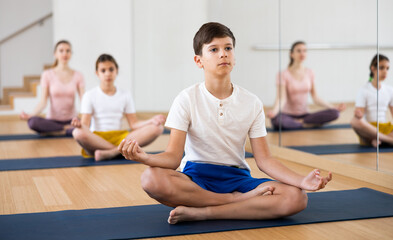 Concentrated teen boy sitting on mat in fitness center, making yoga meditation in lotus pose while exercising with family..